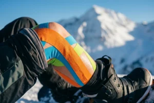 A snowboarder applying synthetic kinesiology tape to their lead knee before hitting the slopes