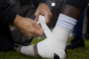 An athletic trainer's hands wrapping a player's ankle using white easy-tear sports tape on a sideline